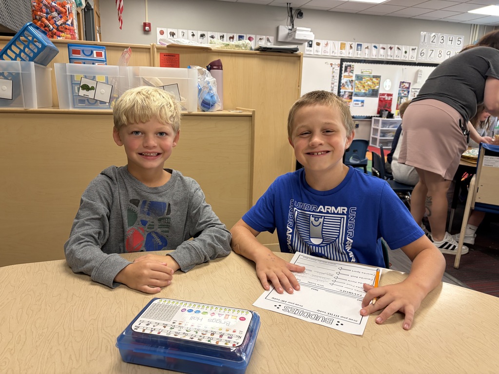 Two young boys sitting together at a table smiling, one wearing a blue Under Armour shirt.