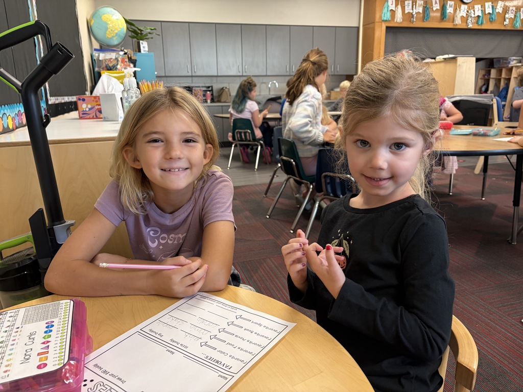 Two smiling girls sitting together at a wooden classroom desk with a worksheet in front of them.