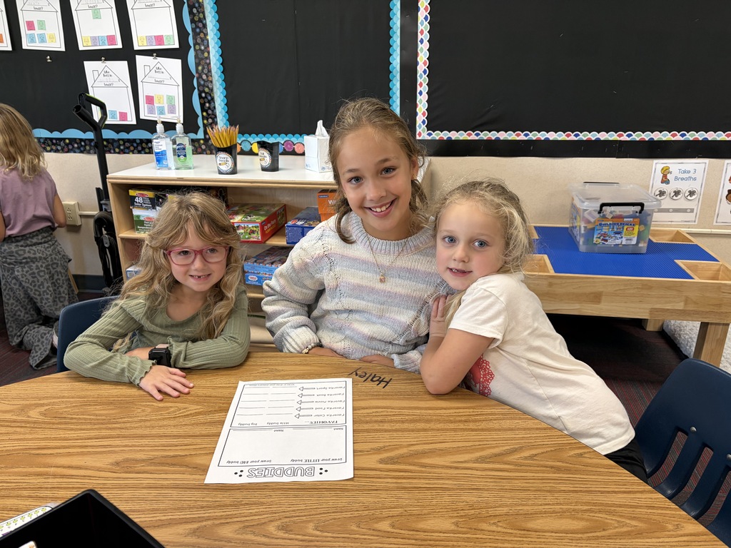 Three young girls seated together around a wooden desk smiling for a group photo.