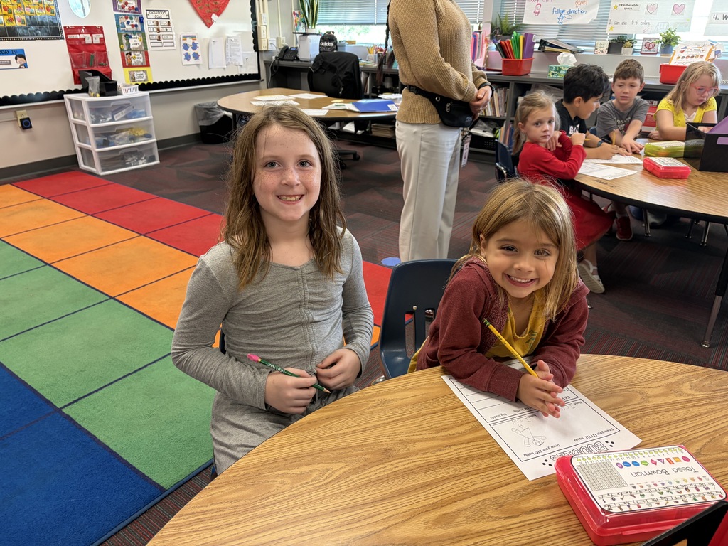 Two smiling young girls sitting together at a classroom desk working on a activity sheet.