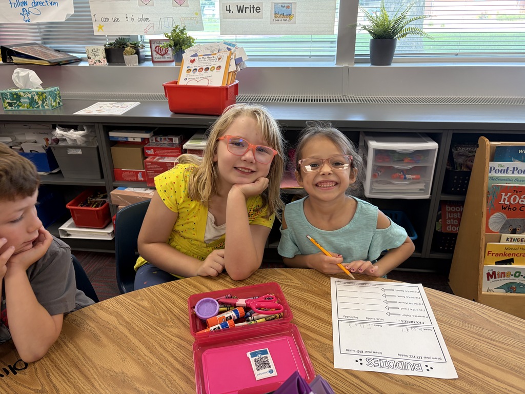 Two young girls with glasses smiling together at a classroom desk with school supplies and worksheets.