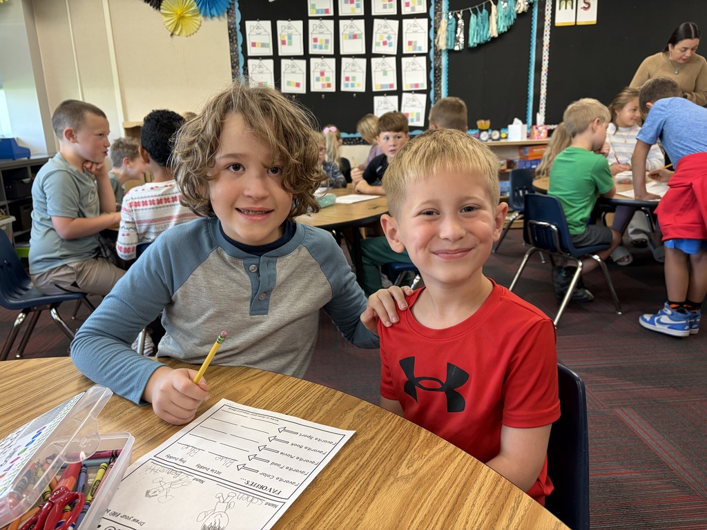 Two young boys smiling at a classroom table while working on a "BUDDIES" activity worksheet.