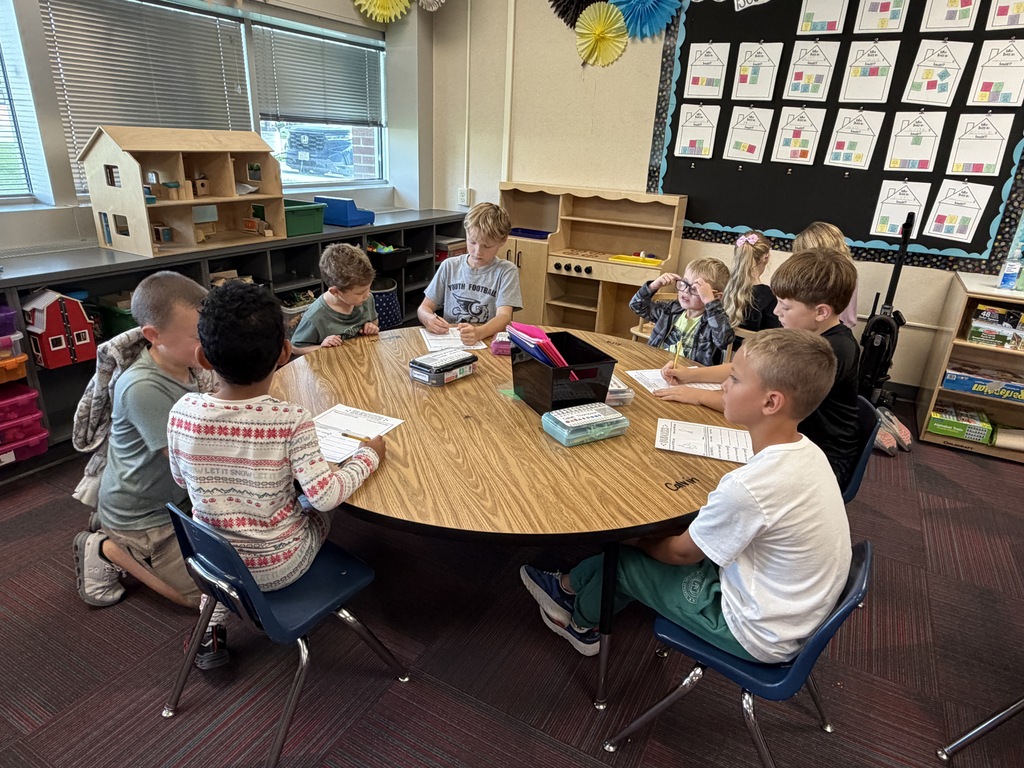 Elementary students seated around a large circular desk working on worksheets together in a classroom.