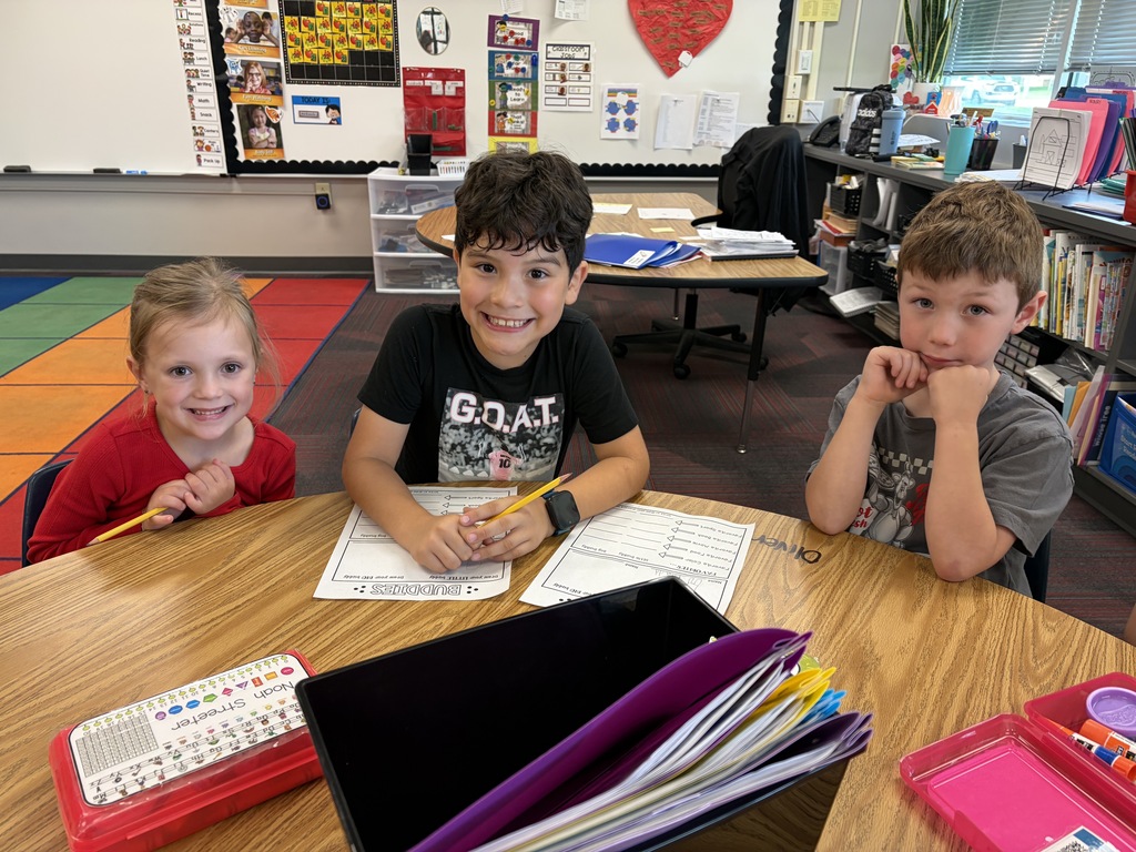 Three elementary students sitting together at a desk with pencils and "BUDDIES" activity sheets.