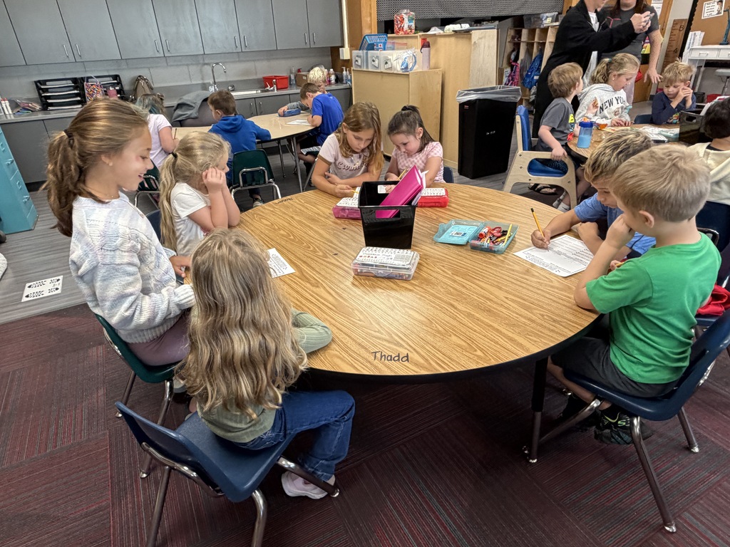 A group of young students sitting around a large round wooden table during classroom instruction time.