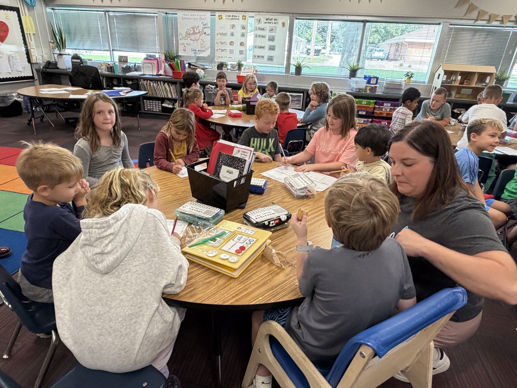 A teacher and several students sitting around a table together with learning materials in a busy classroom.