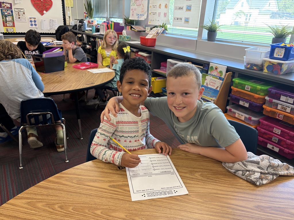 A young boy in a patterned sweater with his arm around another boy smiling at a classroom table.