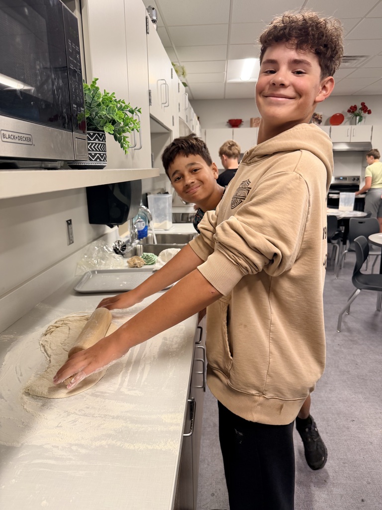 Two students smile at the camera while rolling out dough. 