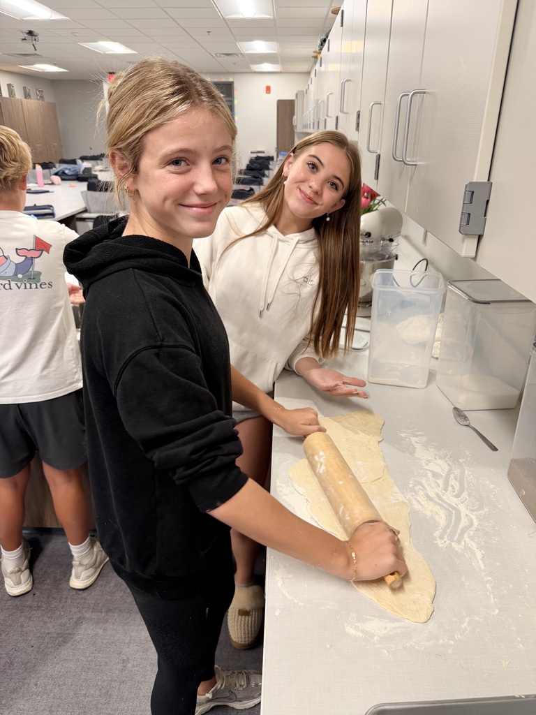 Two students smile at the camera while rolling out dough. 