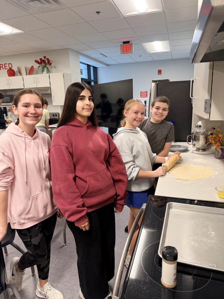 A group of students smile at the camera while rolling out dough. 
