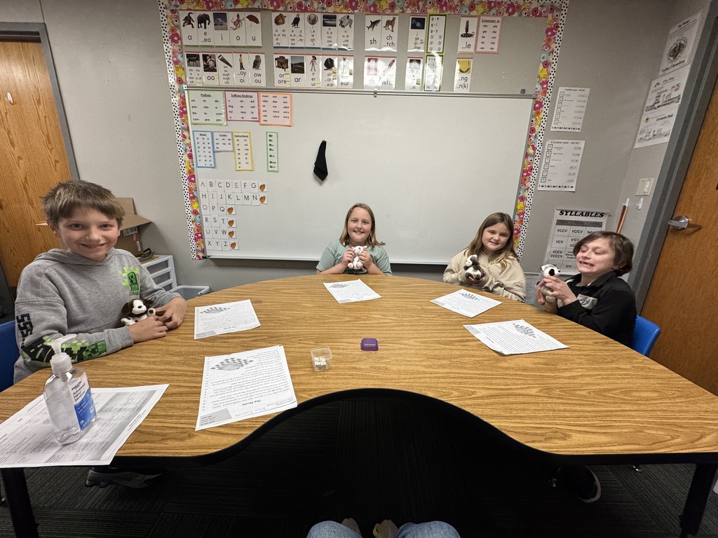 Four students sitting around a kidney-shaped wooden table holding plush animal finger puppets while working on reading worksheets.