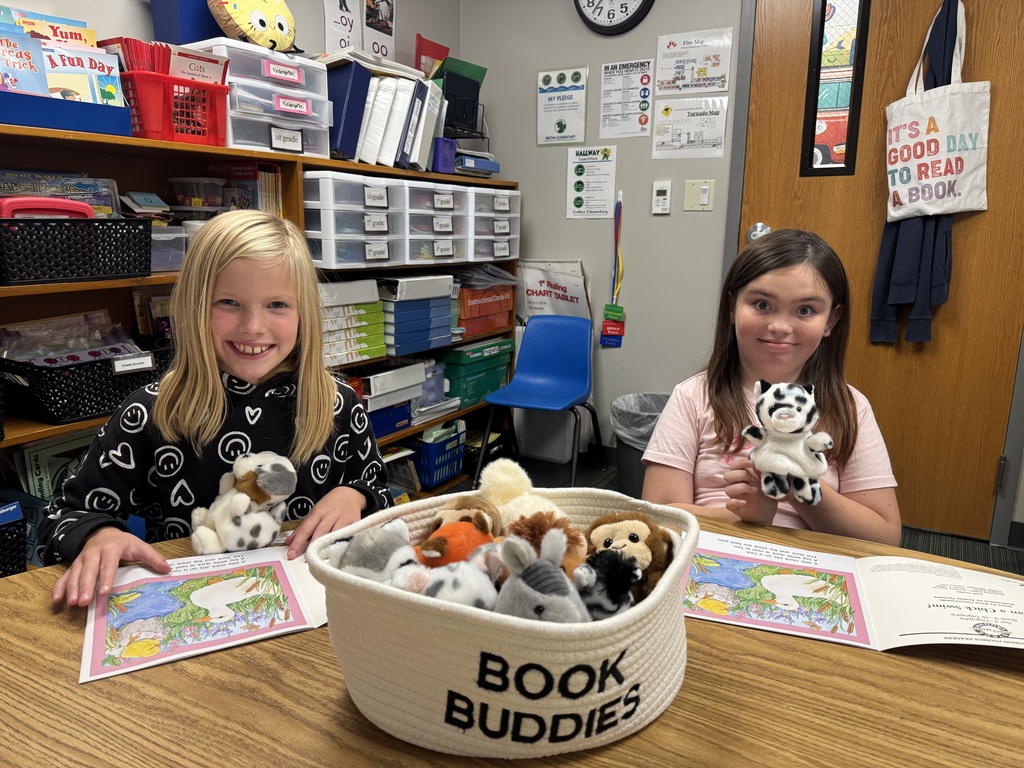 Two smiling girls sitting at a table with open picture books and plush finger puppets, next to a "BOOK BUDDIES" basket filled with stuffed animals.
