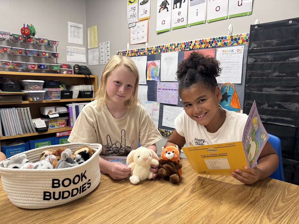 Two young girls smiling at a table while holding small plush animal toys next to an open book and a "BOOK BUDDIES" basket.