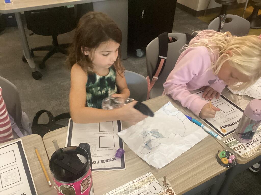 students spraying water on paper for a science project. They are seated at their desk.
