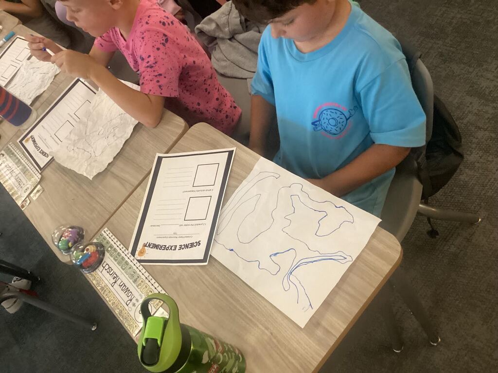 students spraying water on paper for a science project. They are seated at their desk.