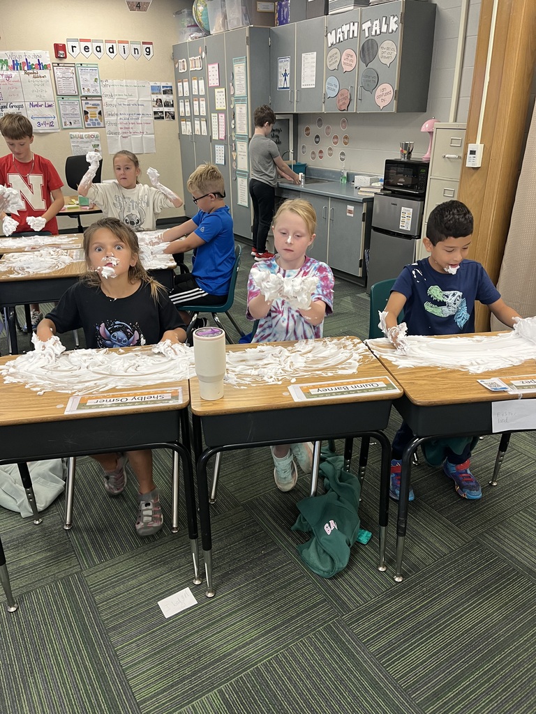 Elementary students sitting at connected classroom desks covered in shaving cream, playfully getting foam on their hands and faces.