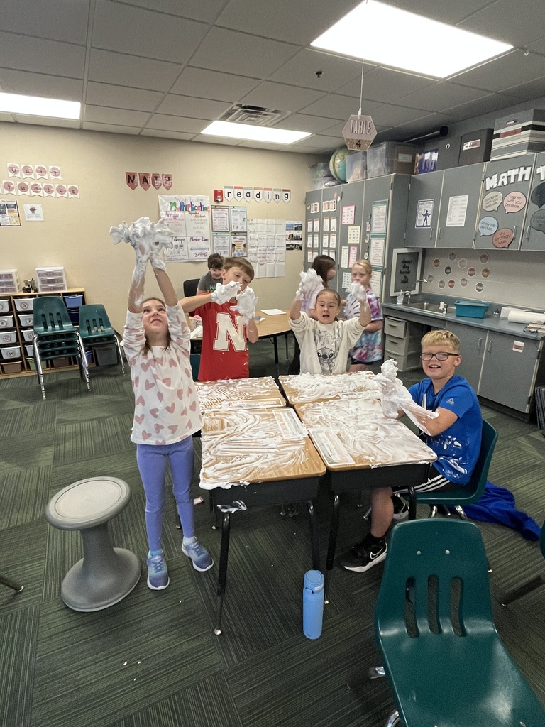 A group of students having fun during a sensory activity, posing and holding up shaving cream on their hands above foam-covered desks.