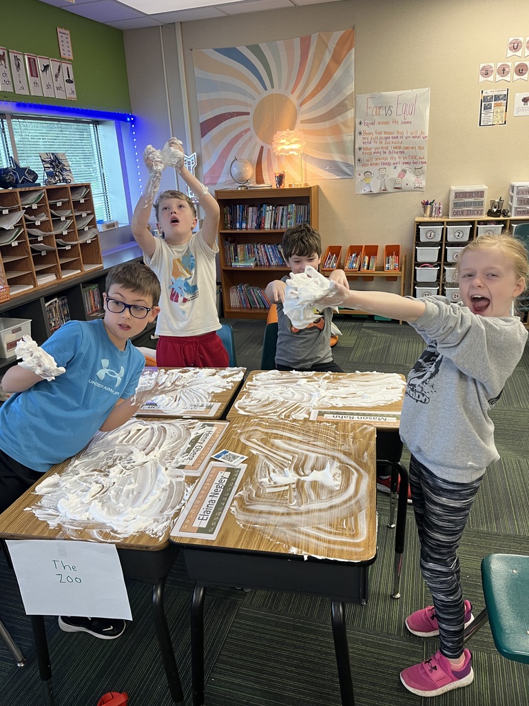 Four young students gathered around desks covered in shaving cream, holding up foam-covered hands and smiling.