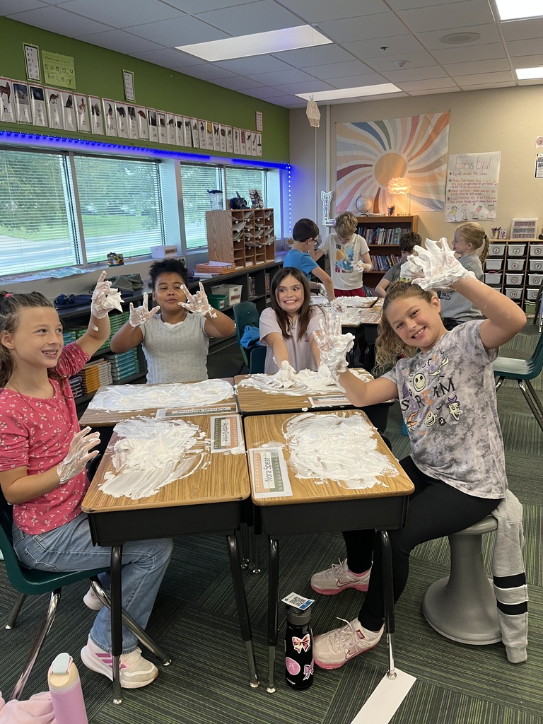 Smiling elementary students showing off foam-covered hands while seated at desks spread with shaving cream.
