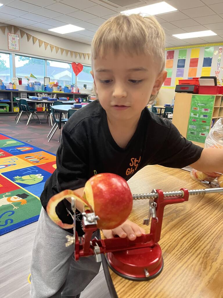A young boy guiding an apple through a red hand-crank apple peeler at a classroom table.