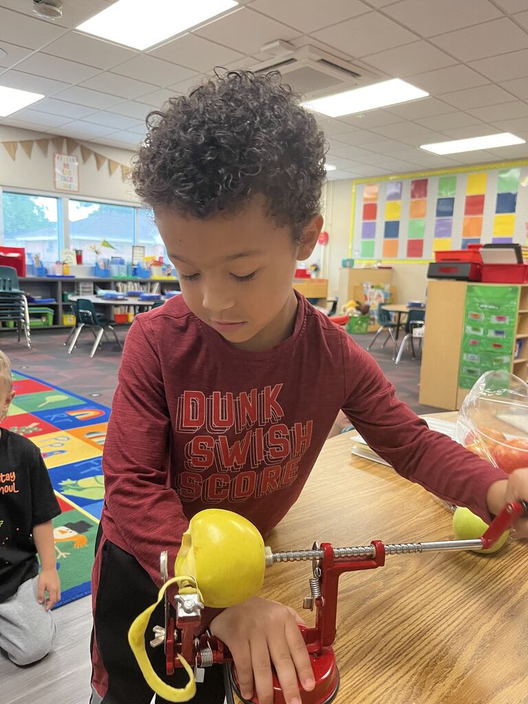 A young boy in a long-sleeved shirt turning the handle on a red hand-crank apple peeler and corer.