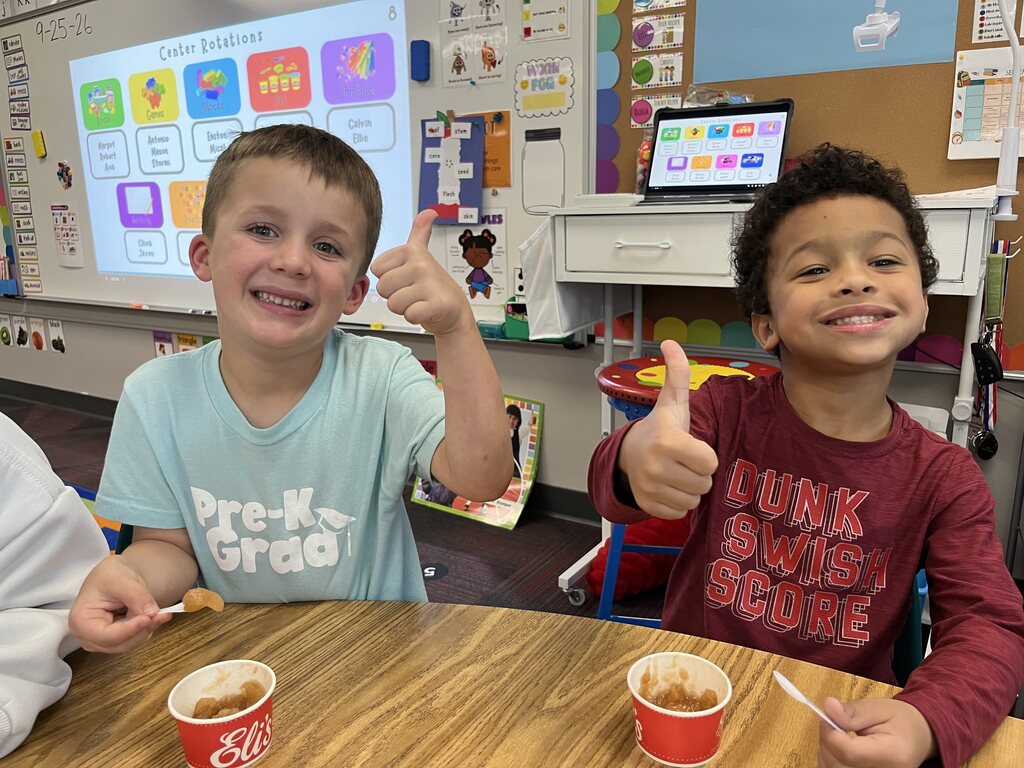 Two young boys giving a thumbs-up while sitting at a desk with cups of fresh applesauce.