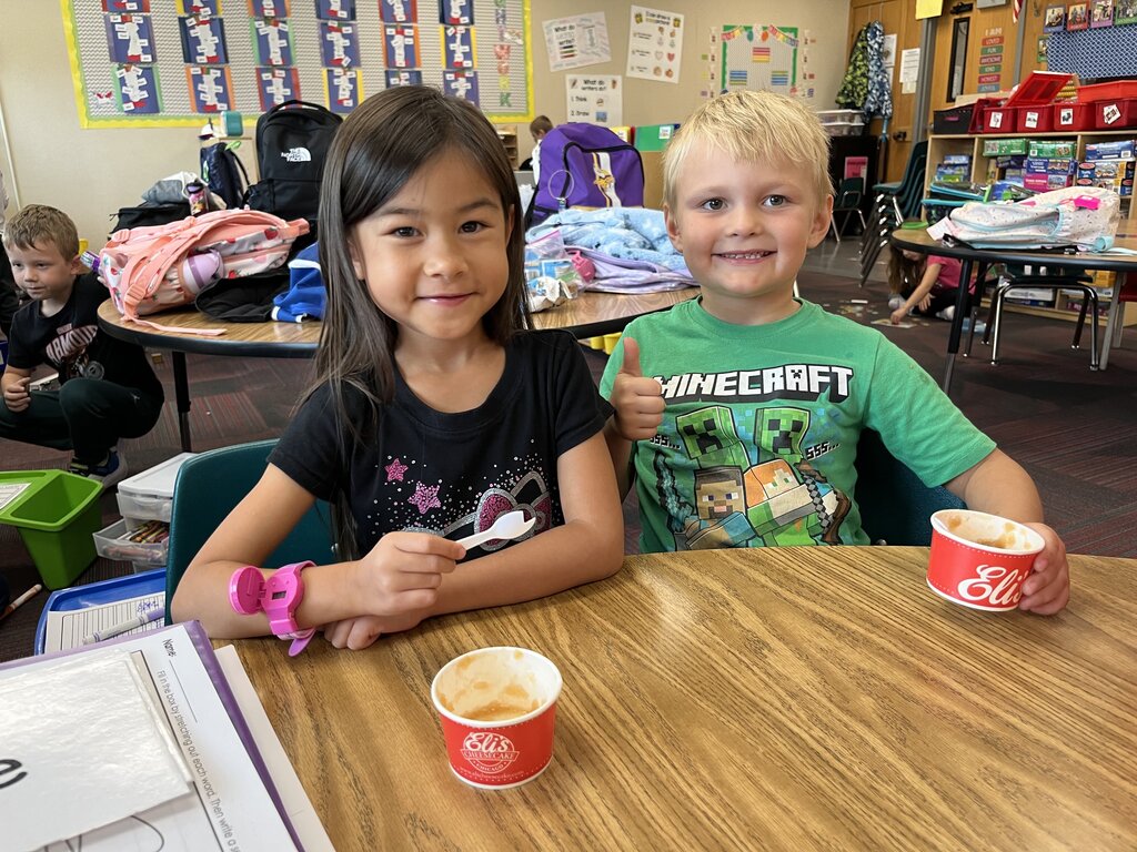 Two students smiling and giving a thumbs-up at a wooden desk with their applesauce cups.