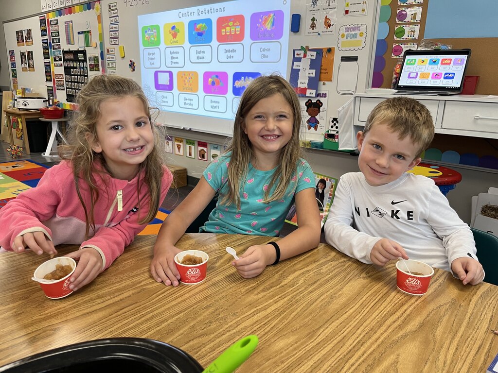 Three young students smiling at a table with their small paper cups of fresh applesauce.