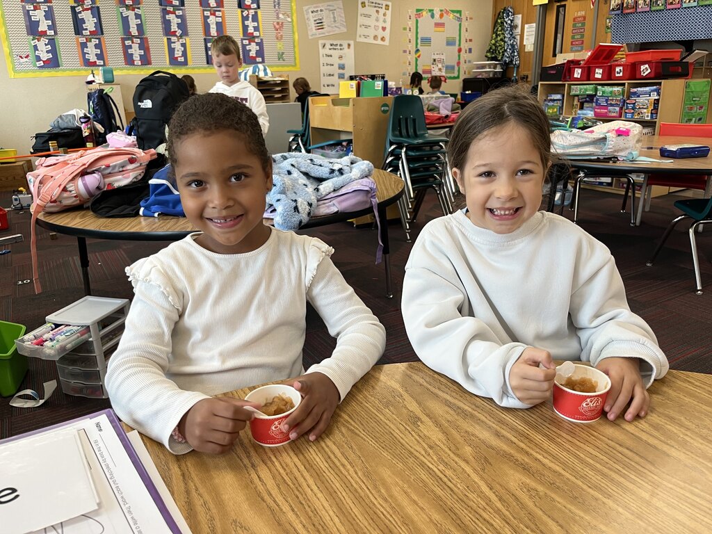 Two smiling students sitting together at a wooden table enjoying small cups of fresh applesauce with plastic spoons.