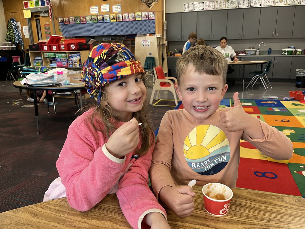 Two smiling young students, one giving a thumbs-up, enjoying fresh applesauce at a classroom table.