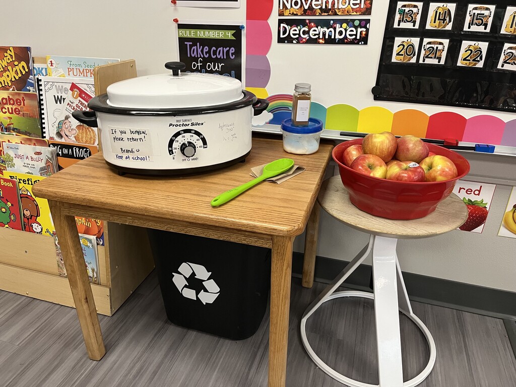 A slow cooker set up on a wooden table next to a red bowl filled with fresh apples and ingredients for making applesauce.