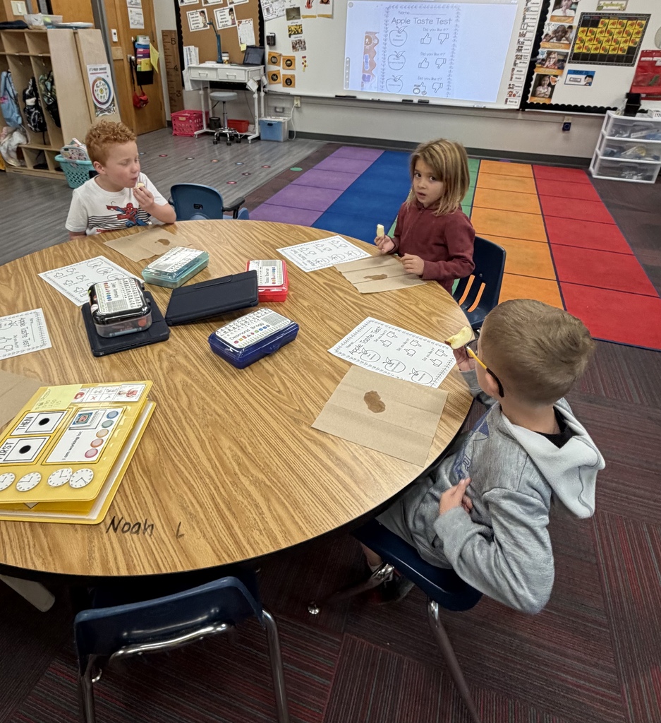 Three students seated around a round wooden table participating in an apple taste test with worksheets and apple slices.
