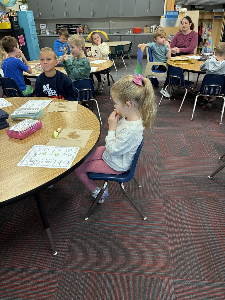 Classroom view of several students seated at round tables sampling different types of apples for a taste test lesson.