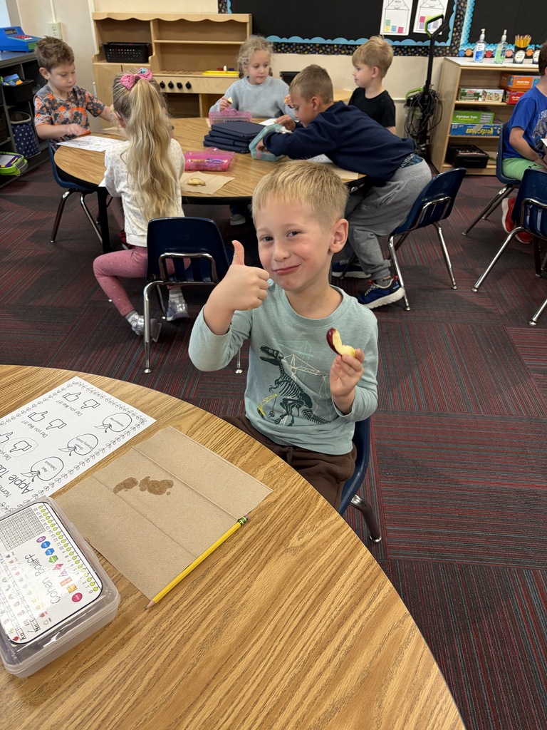 A young boy holding up an apple slice and giving a thumbs-up during a classroom apple taste test activity.