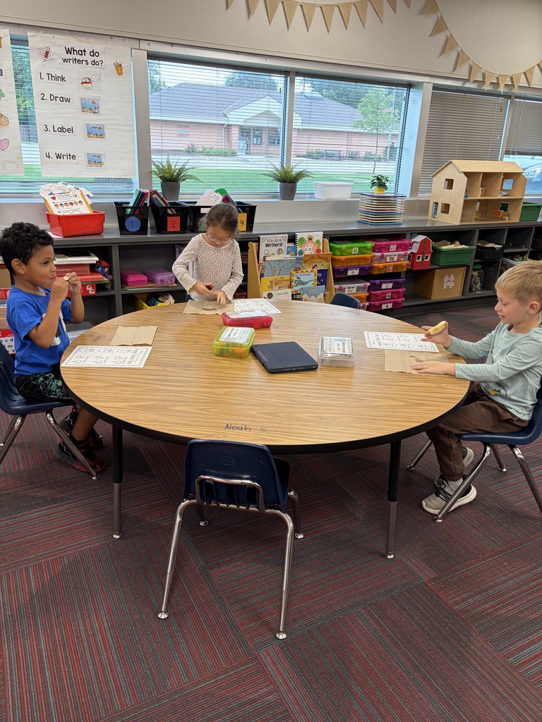 Three young students sitting at a round wooden table in a classroom with "Apple Taste Test" worksheets in front of them.