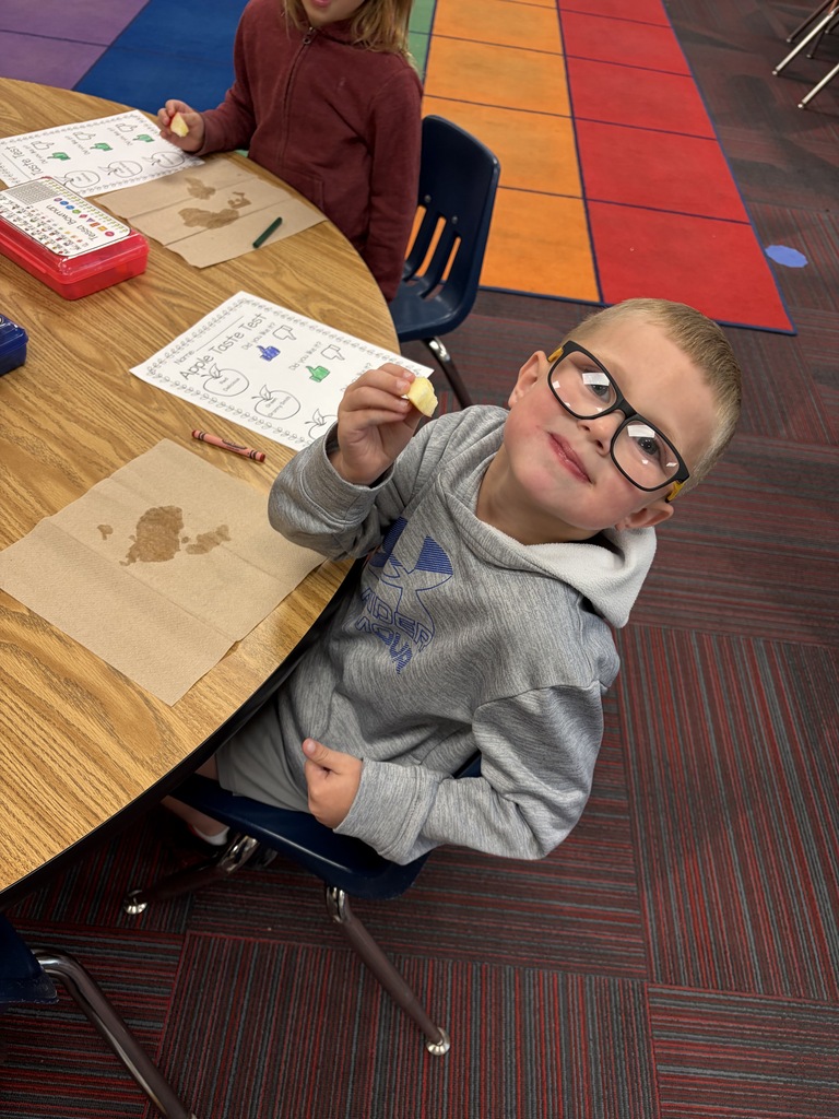 A boy with glasses holding up a slice of apple to taste during a classroom lesson.