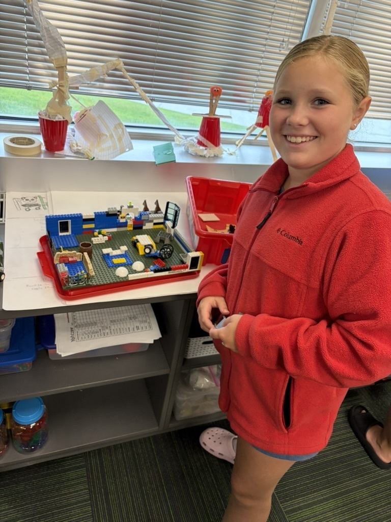 A student standing proudly in her red fleece jacket next to a detailed Lego build displayed on a tray.