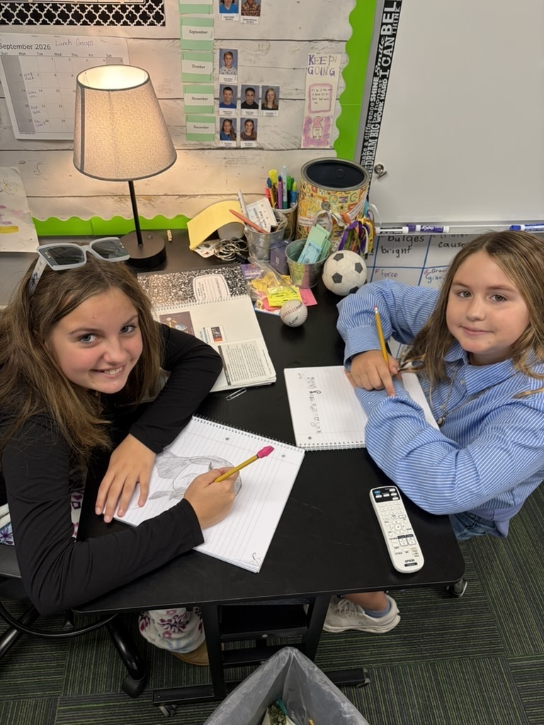 Two female students working together at a desk on drawing and writing activities next to a desk lamp.