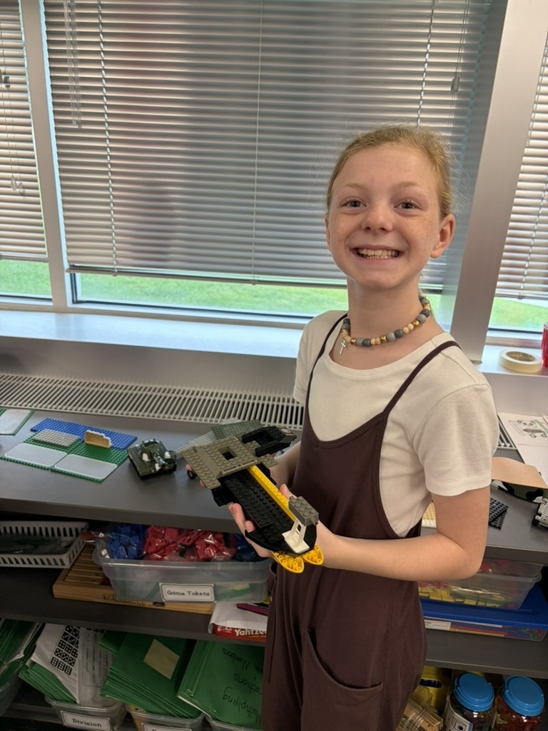 A smiling girl wearing overall pants showing off her custom Lego creation in the classroom.