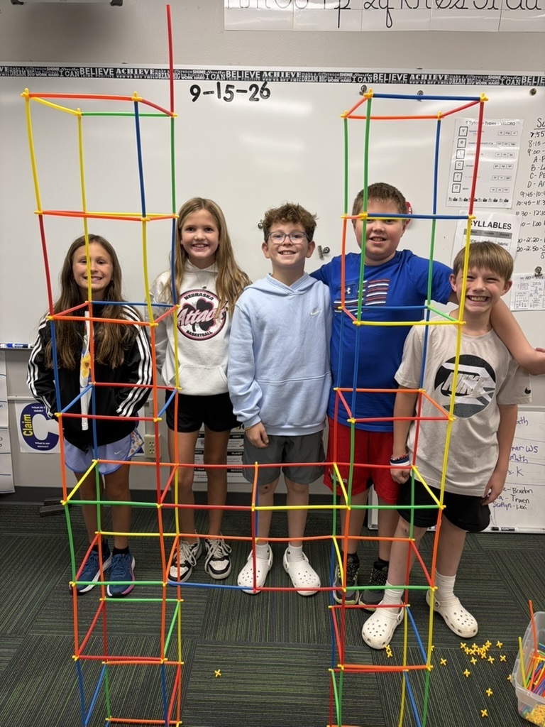 Five smiling elementary students posing behind a large, colorful straw and connector tower structure built in their classroom.