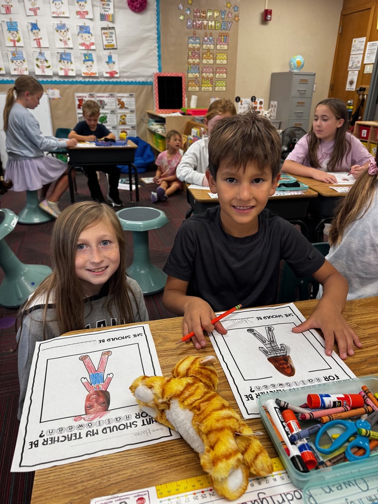 A girl and a boy smiling at their desk while working on Halloween-themed art worksheets alongside a small stuffed lion toy.