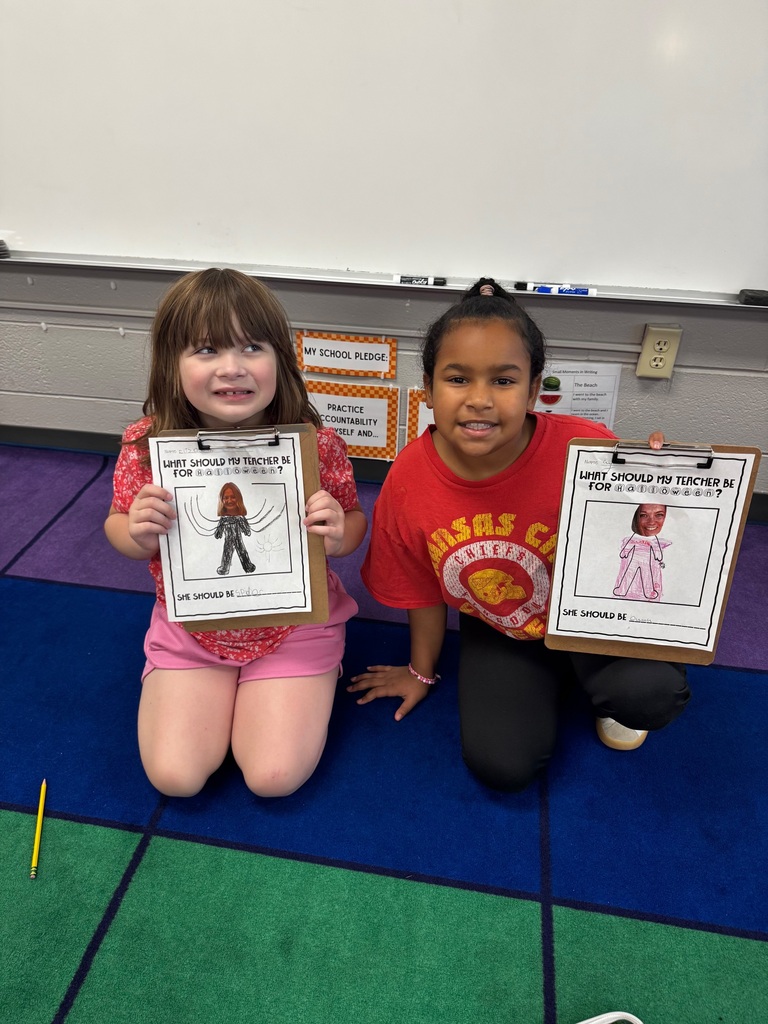 Two young girls sitting on a classroom carpet holding clipboard activity sheets with costume ideas for their teacher.