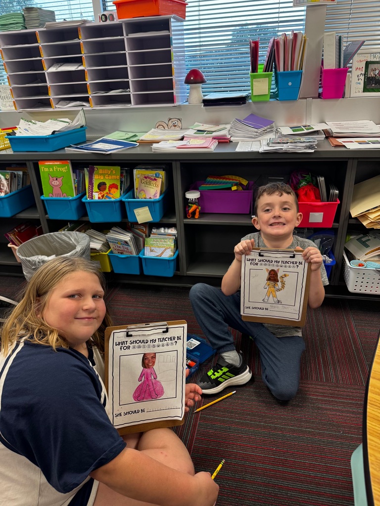 Two students holding up clipboards featuring colorful teacher Halloween costume illustrations in front of classroom bookshelf cubbies.