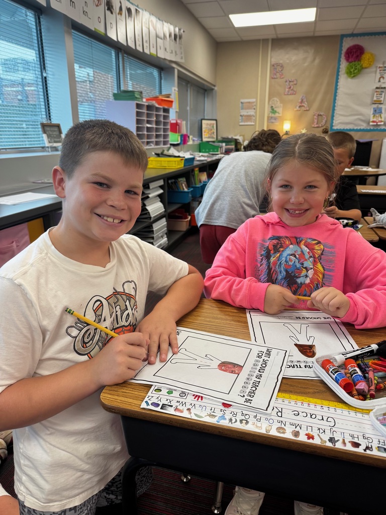Two elementary students smiling at their desk while filling out a Halloween worksheet titled "What Should My Teacher Be For Halloween?".