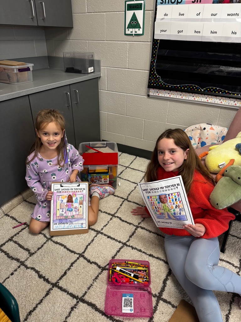 Two young girls sitting on a patterned rug in the corner of a classroom displaying their finished art projects.