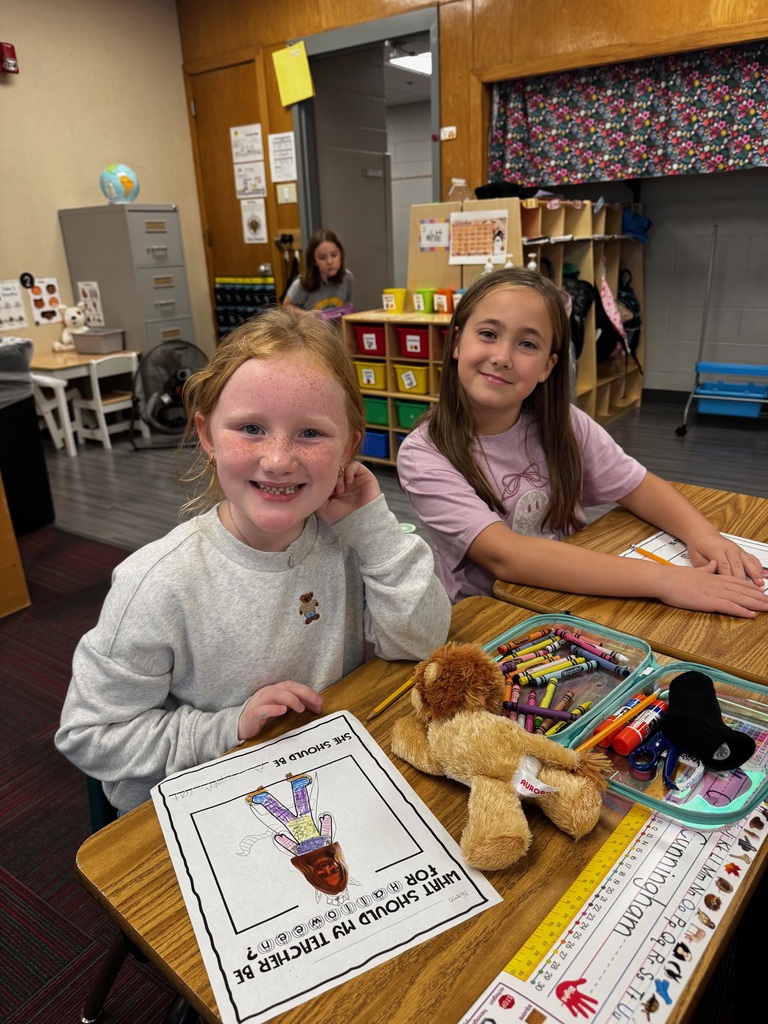 Two elementary girls seated together at a desk smiling alongside a stuffed animal and art supplies.