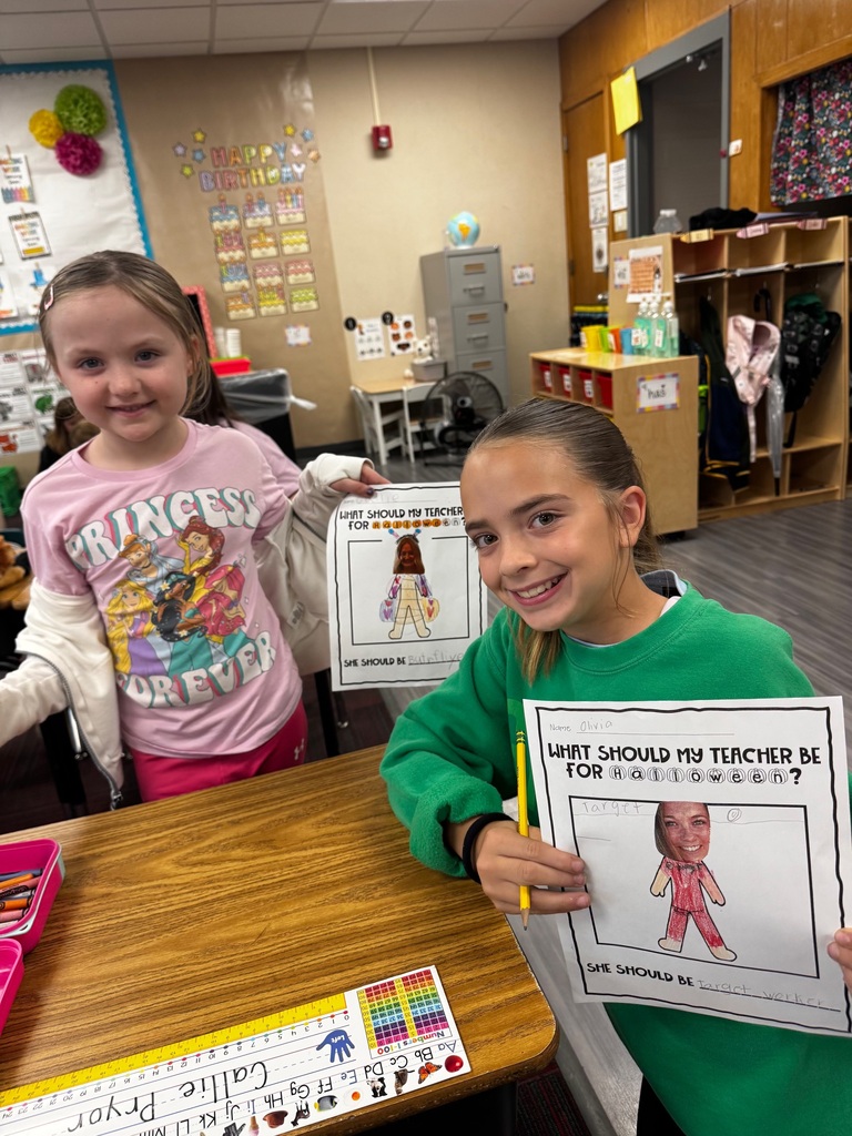 Two smiling girls sitting together at a desk displaying their completed "What Should My Teacher Be For Halloween?" pages.
