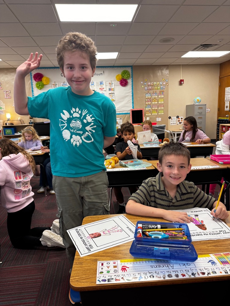 A boy standing and waving next to a seated classmate working on a Halloween drawing worksheet at his desk.