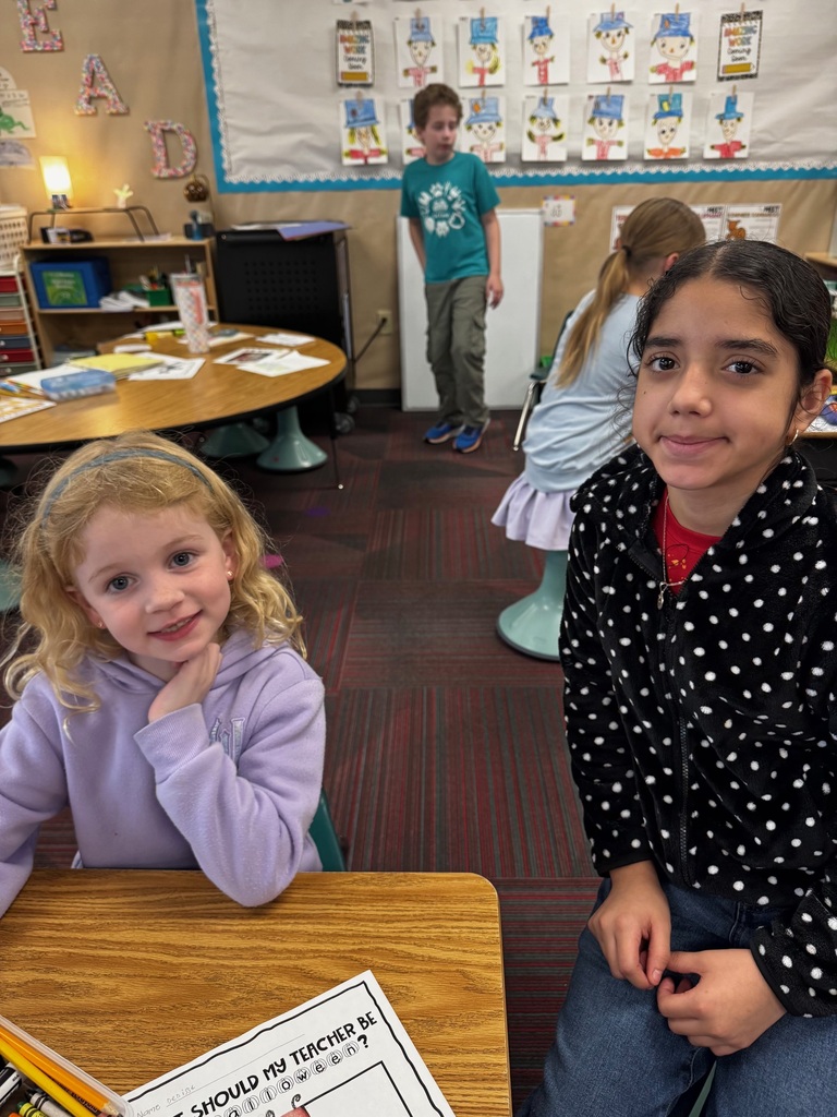Two girls posing at a wooden classroom desk while participating in a creative drawing activity.