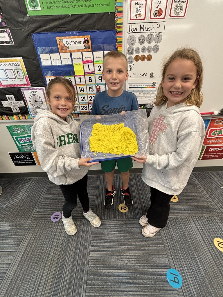 Three elementary students holding a tray displaying their bright yellow play dough structure in front of a classroom calendar.